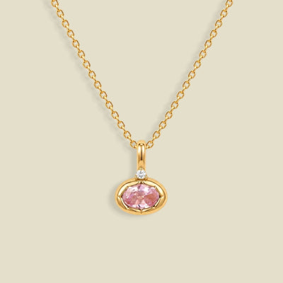 A delicate gold chain necklace with a small oval pendant featuring a pink gemstone and a tiny clear accent stone above it, set against a solid light beige background.