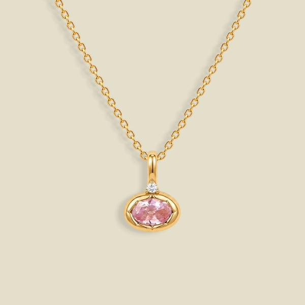 A delicate gold chain necklace with a small oval pendant featuring a pink gemstone and a tiny clear accent stone above it, set against a solid light beige background.