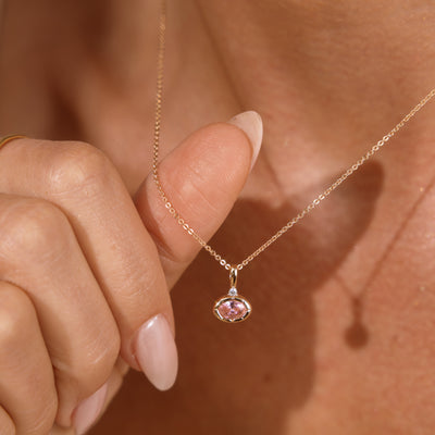 A close-up lifestyle shot of a delicate gold chain necklace with a small oval pink bezel-set gemstone pendant, worn on a model's neck and held by a hand with manicured nails.