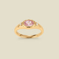 A gold signet ring with a pink center oval bezel-set gemstone, and two small clear accent stones on either side, set against a solid beige background.