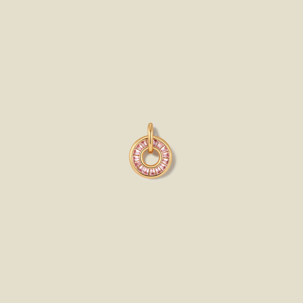 A circular gold pendant featuring pink baguette-cut stones around a hollow center, displayed against a solid light beige background.
