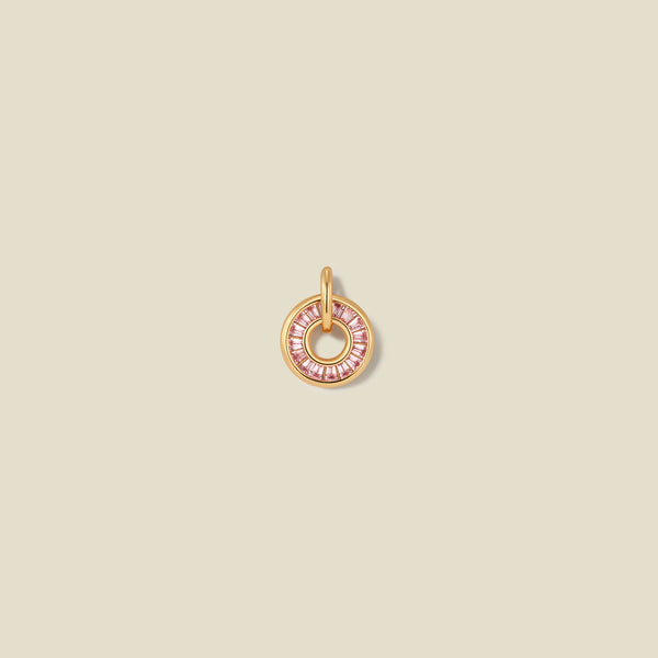 A circular gold pendant featuring pink baguette-cut stones around a hollow center, displayed against a solid light beige background.