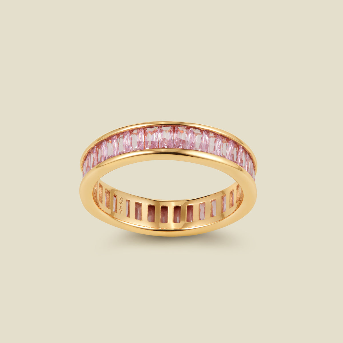 A gold eternity ring featuring a continuous channel setting of baguette-cut pink gemstones, shown on a solid light beige background.