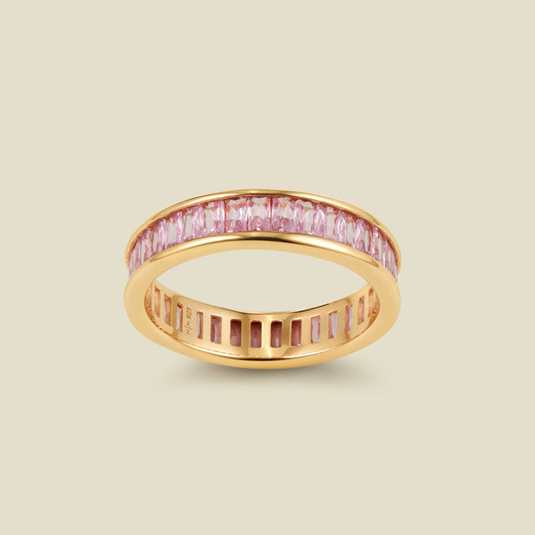 A gold eternity ring featuring a continuous channel setting of baguette-cut pink gemstones, shown on a solid light beige background.