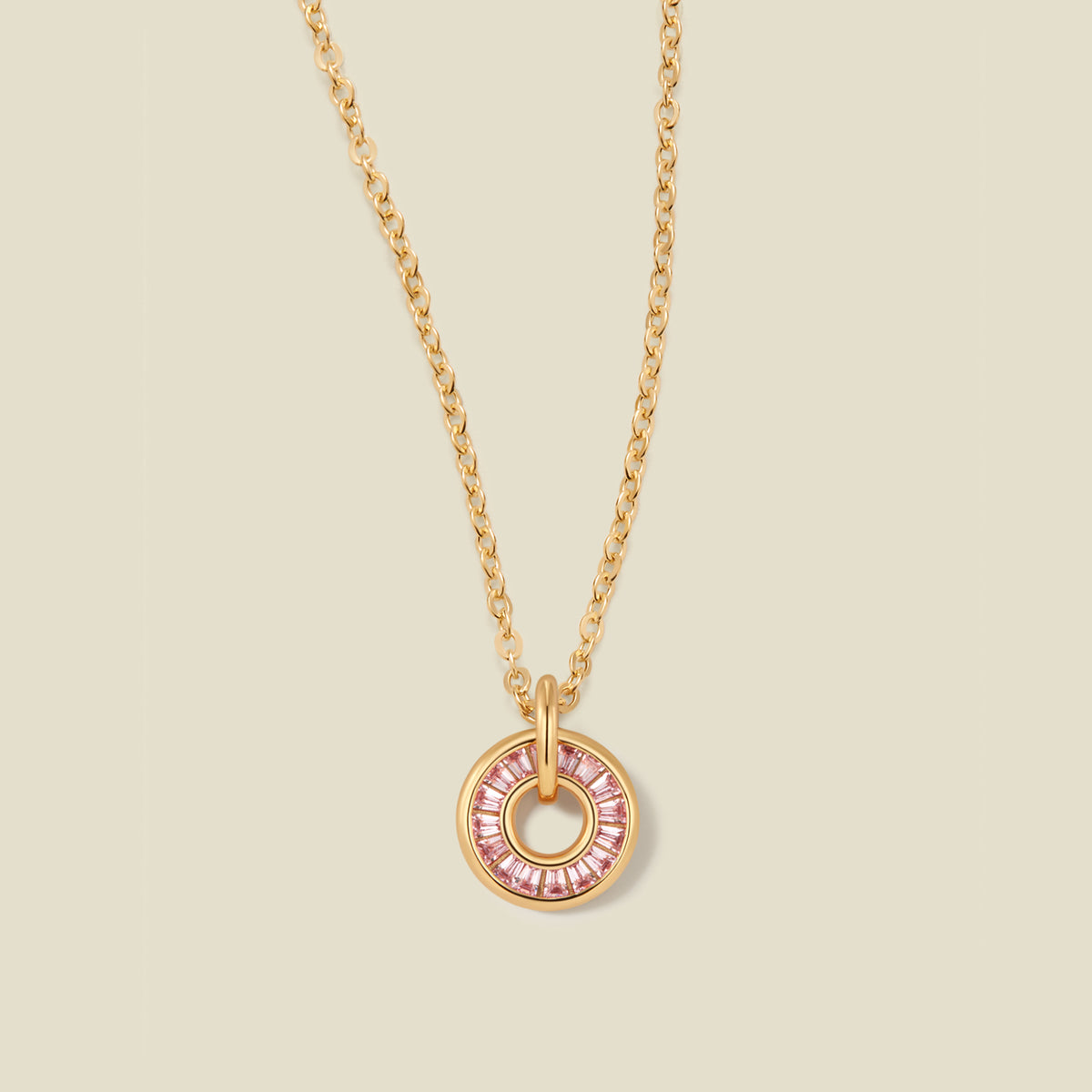 A gold chain necklace with a circular pendant featuring pink baguette-cut stones around a hollow center, displayed against a solid light beige background.