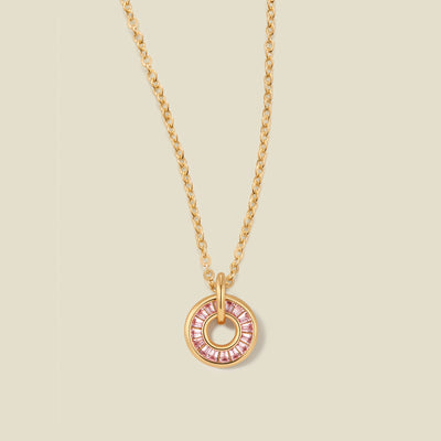 A gold chain necklace with a circular pendant featuring pink baguette-cut stones around a hollow center, displayed against a solid light beige background.