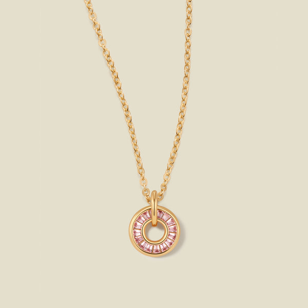 A gold chain necklace with a circular pendant featuring pink baguette-cut stones around a hollow center, displayed against a solid light beige background.