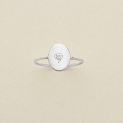 A silver ring with an oval face engraved with a poppy flower, centered on a textured beige background.
