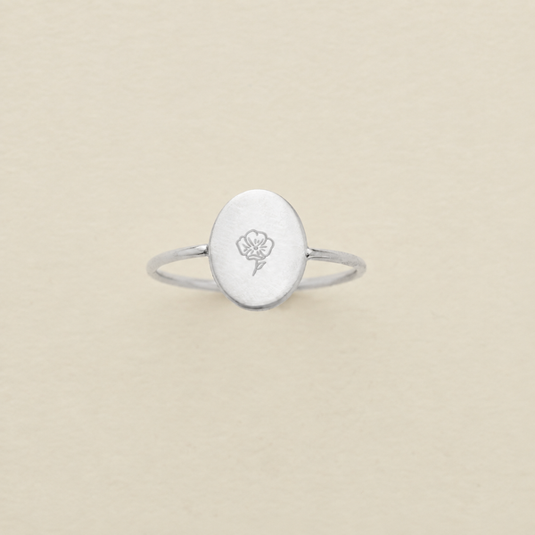 A silver ring with an oval face engraved with a poppy flower, centered on a textured beige background.