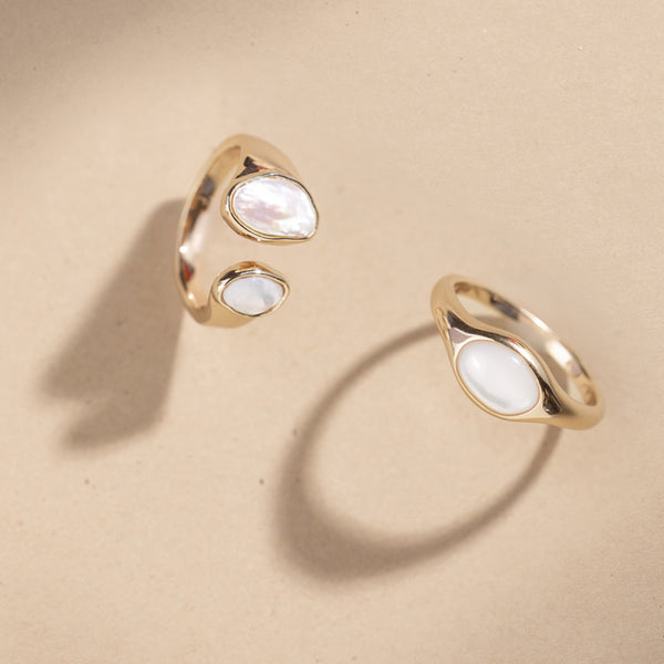 Two gold bezel-set pearl rings against a plain beige background.