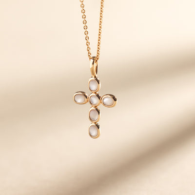Close-up of a gold necklace with a cross pendant made of bezel-set pearl stones, suspended against a neutral background.