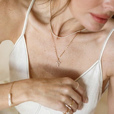Close-up of a person wearing layered gold and pearl necklaces, including a small cross pendant with bezel-set pearls, along with a pearl bracelet and gold rings.