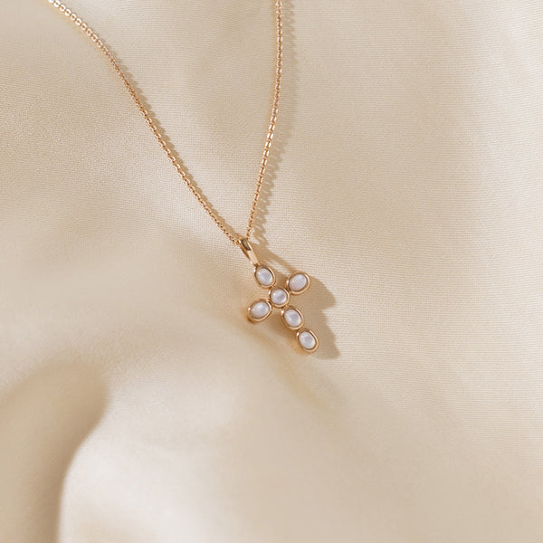 Gold chain and a cross pendant with bezel-set pearls, on a soft fabric backdrop.