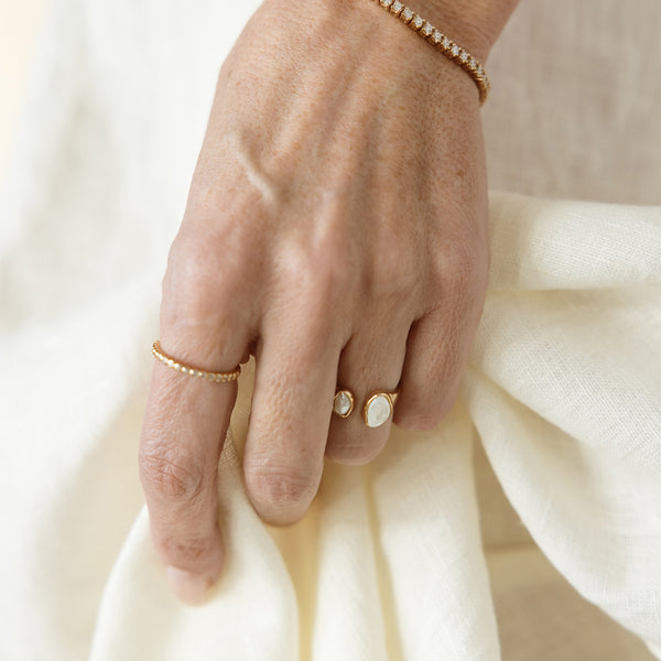 Close-up of a hand wearing multiple delicate gold rings with bezel-set pearl stones and a tennis bracelet resting on soft fabric.