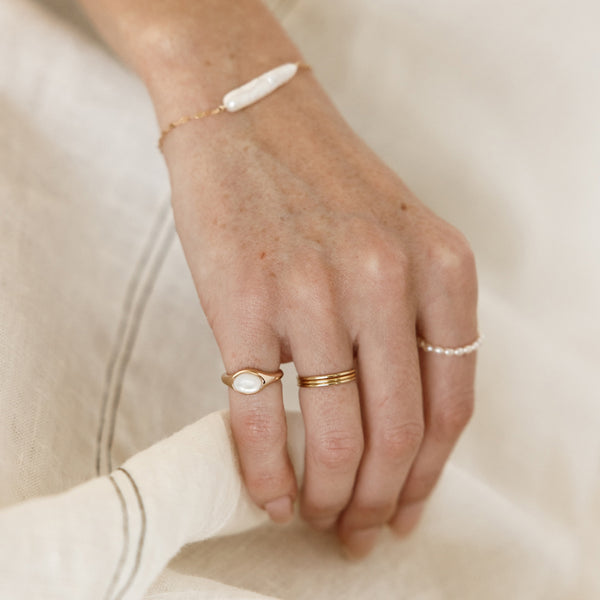 Hand lightly touching linen wearing gold stacked rings, a bezel-set pearl pinky ring and a baroque pearl bracelet.