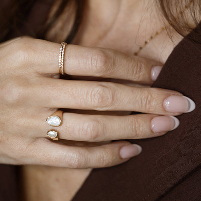 Close-up of a hand wearing gold rings, including a double open ring with bezel-set pearl stones and a thin stacked band, resting against a brown shirt.