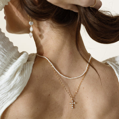 Woman adjusting her hair while wearing layered necklaces, including a delicate pearl strand and a gold chain with a cross pendant featuring bezel-set pearls, paired with drop pearl earrings.