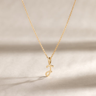 A delicate gold chain necklace with a small cursive initial "J" pendant, shown against a soft beige background with natural shadows.