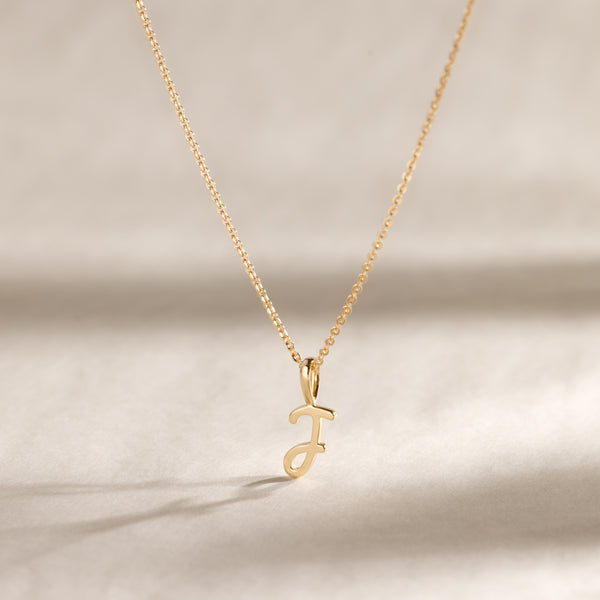 A delicate gold chain necklace with a small cursive initial "J" pendant, shown against a soft beige background with natural shadows.