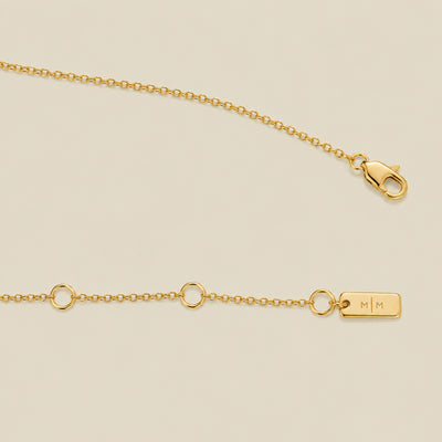 A gold chain with a lobster clasp, small circular links, and a rectangular engraved tag marked 'M|M', shown on a light beige background.