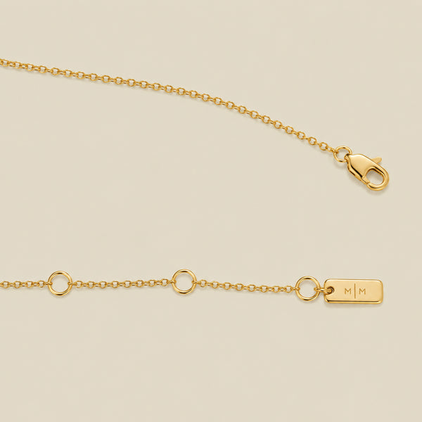 A gold chain with a lobster clasp, small circular links, and a rectangular engraved tag marked 'M|M', shown on a light beige background.