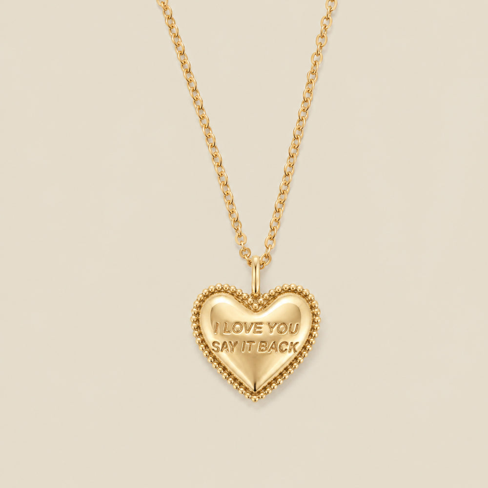 A gold heart-shaped pendant necklace with a beaded border and the engraved words 'I LOVE YOU SAY IT BACK', hanging from a gold chain against a solid beige background.