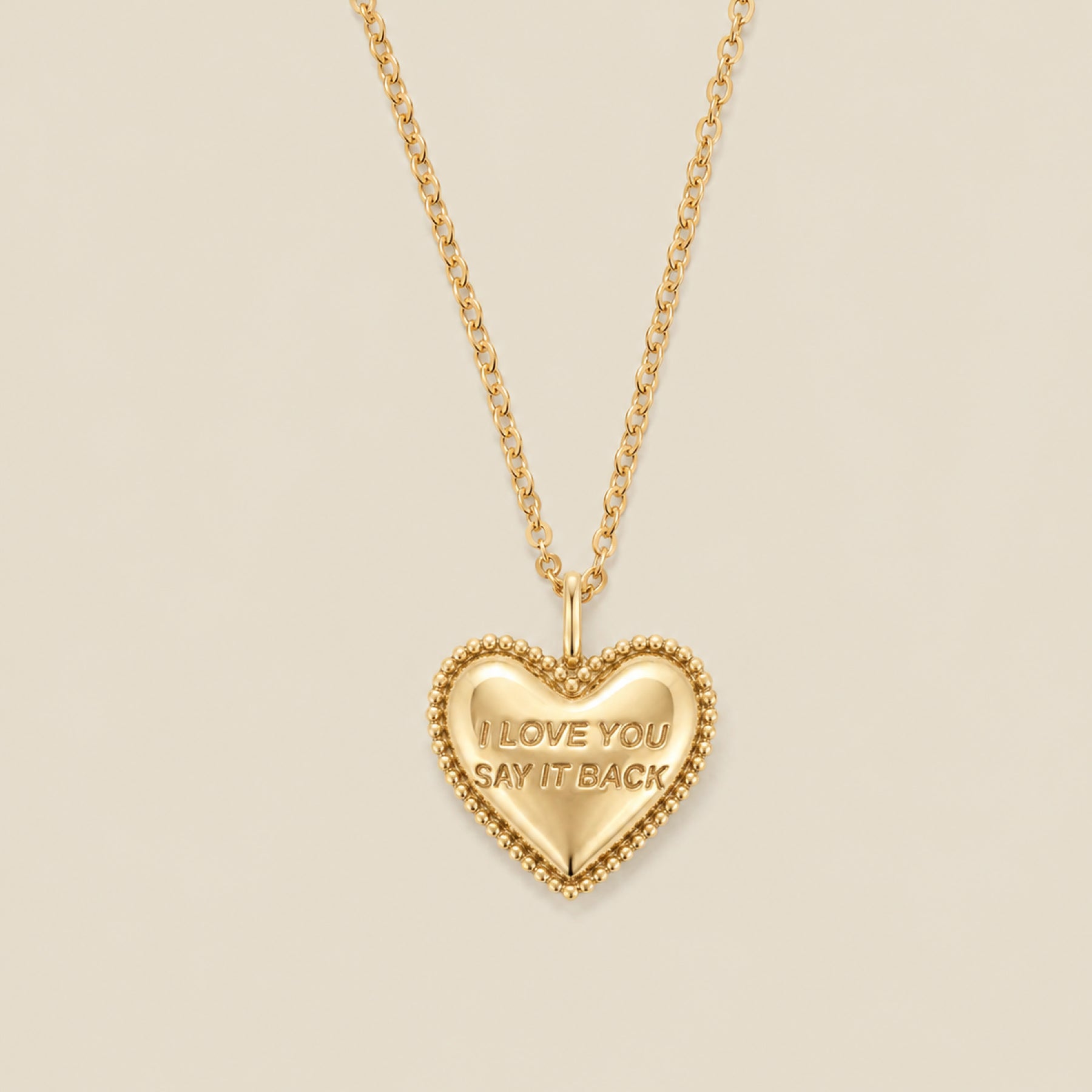 A gold heart-shaped pendant necklace with a beaded border and the engraved words 'I LOVE YOU SAY IT BACK', hanging from a gold chain against a solid beige background.