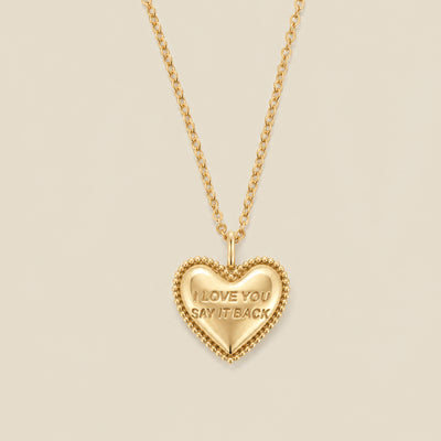 A gold heart-shaped pendant necklace with a beaded border and the engraved words 'I LOVE YOU SAY IT BACK', hanging from a gold chain against a solid beige background.