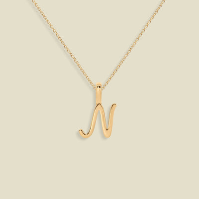 A gold initial necklace featuring a cursive uppercase letter 'N' pendant on a delicate gold chain, set against a solid light beige background.