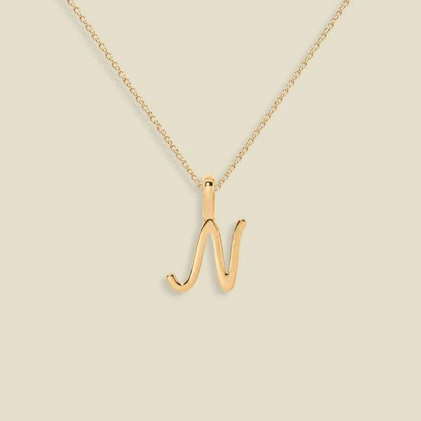 A gold initial necklace featuring a cursive uppercase letter 'N' pendant on a delicate gold chain, set against a solid light beige background.