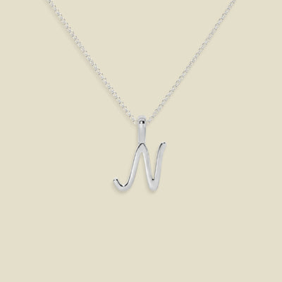 A silver initial necklace featuring a polished uppercase letter 'N' pendant on a delicate silver chain, set against a solid light beige background.