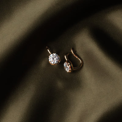 A pair of small gold hoop earrings with clear gemstones, resting on draped dark olive green fabric.
