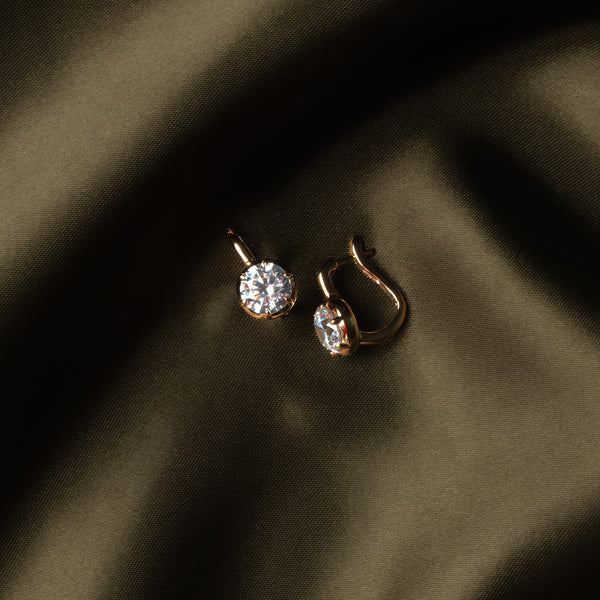 A pair of small gold hoop earrings with clear gemstones, resting on draped dark olive green fabric.