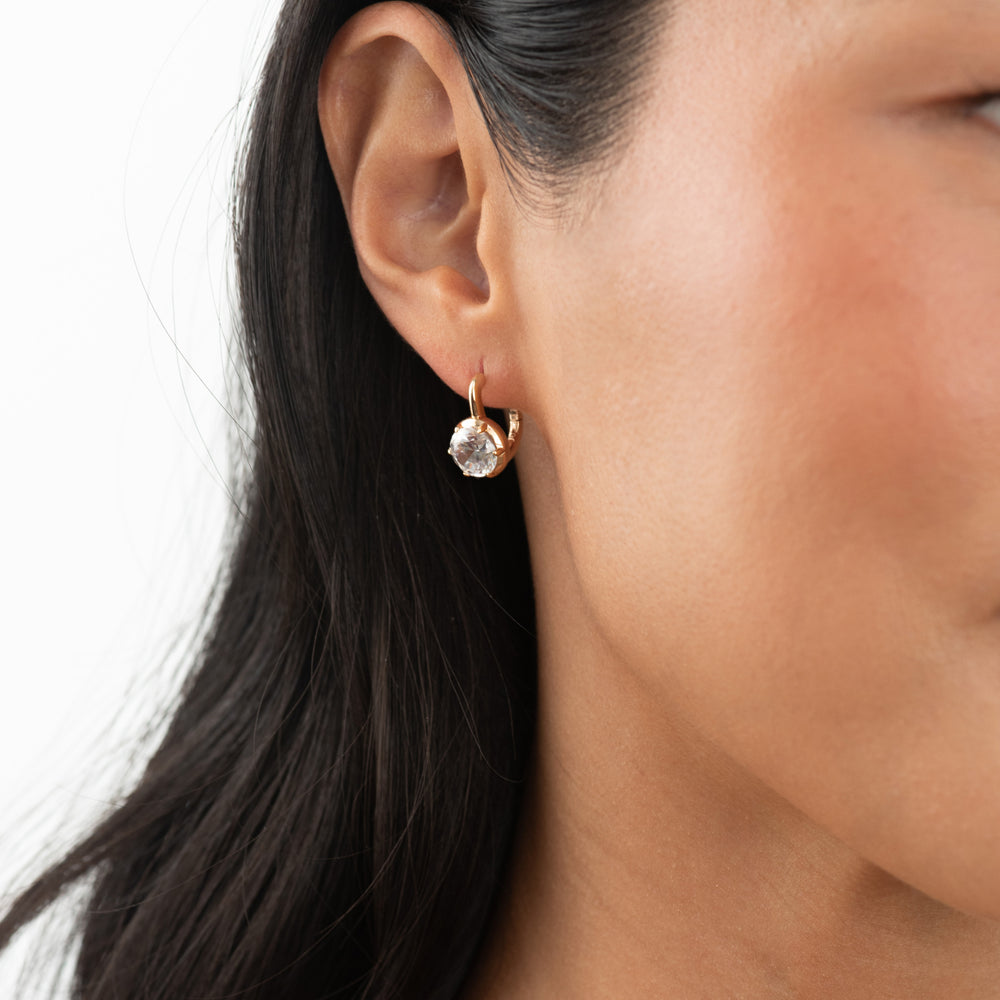 A close-up of a model's ear wearing a small gold hoop earring with a clear faceted gemstone.