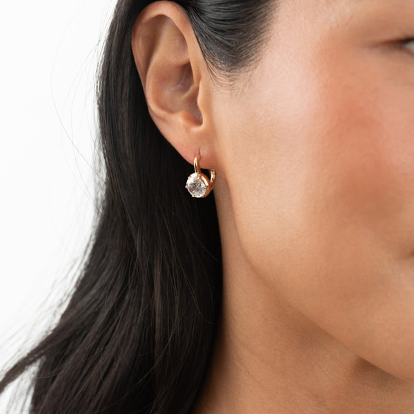 A close-up of a model's ear wearing a small gold hoop earring with a clear faceted gemstone.