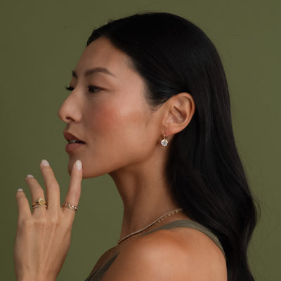 A woman in profile wearing a small gold round drop stud earring, with matching gold rings on her fingers, set against a solid olive green background.