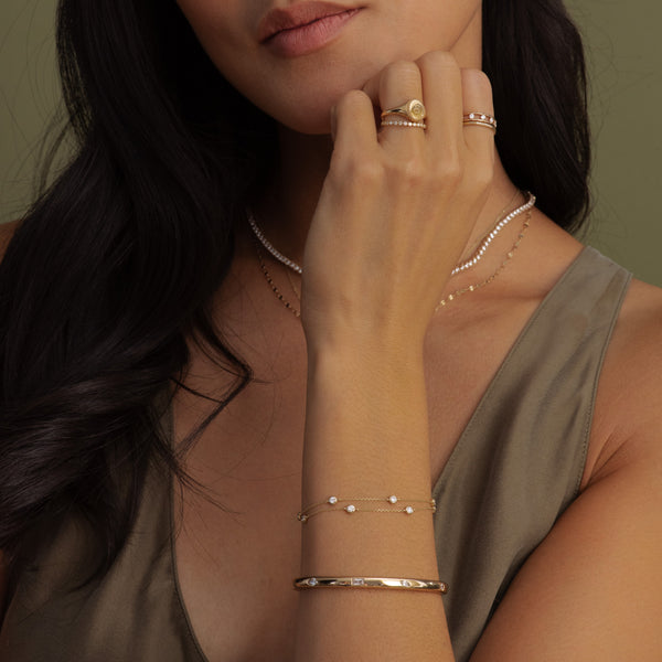 A close-up lifestyle shot of a woman wearing delicate gold jewelry, including a gold bangle bracelet, rings, and layered necklaces, paired with an olive green top against a soft olive green background.