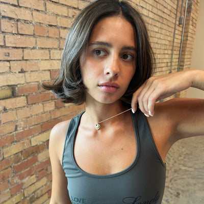 A woman with shoulder-length brown hair wearing a dark gray athletic tank top and a delicate gold heart necklace, posing in front of a brick wall.