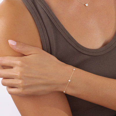 A delicate gold chain bracelet with three small round-shaped settings with clear diamonds worn on a model's wrist, paired with a ribbed brown tank top.