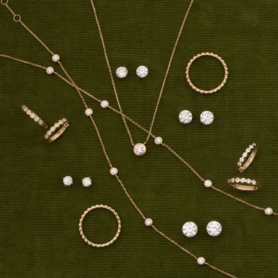 A styled flat-lay of gold jewelry featuring necklaces, rings, and stud earrings with clear gemstones and diamond accents, arranged on a textured dark olive green background.