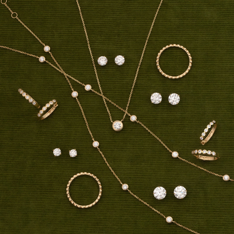A styled flat-lay of gold jewelry featuring necklaces, rings, and stud earrings with clear gemstones and diamond accents, arranged on a textured dark olive green background.