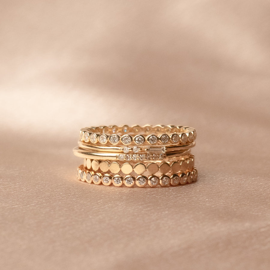 A stack of multiple gold rings, some with clear gemstones in different styles and patterns, set against a cream colored satin background.