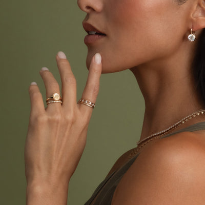 A close-up of a woman's profile resting her hand on her chin, showcasing her hand with multiple gold rings in different styles, set against an olive green backdrop.