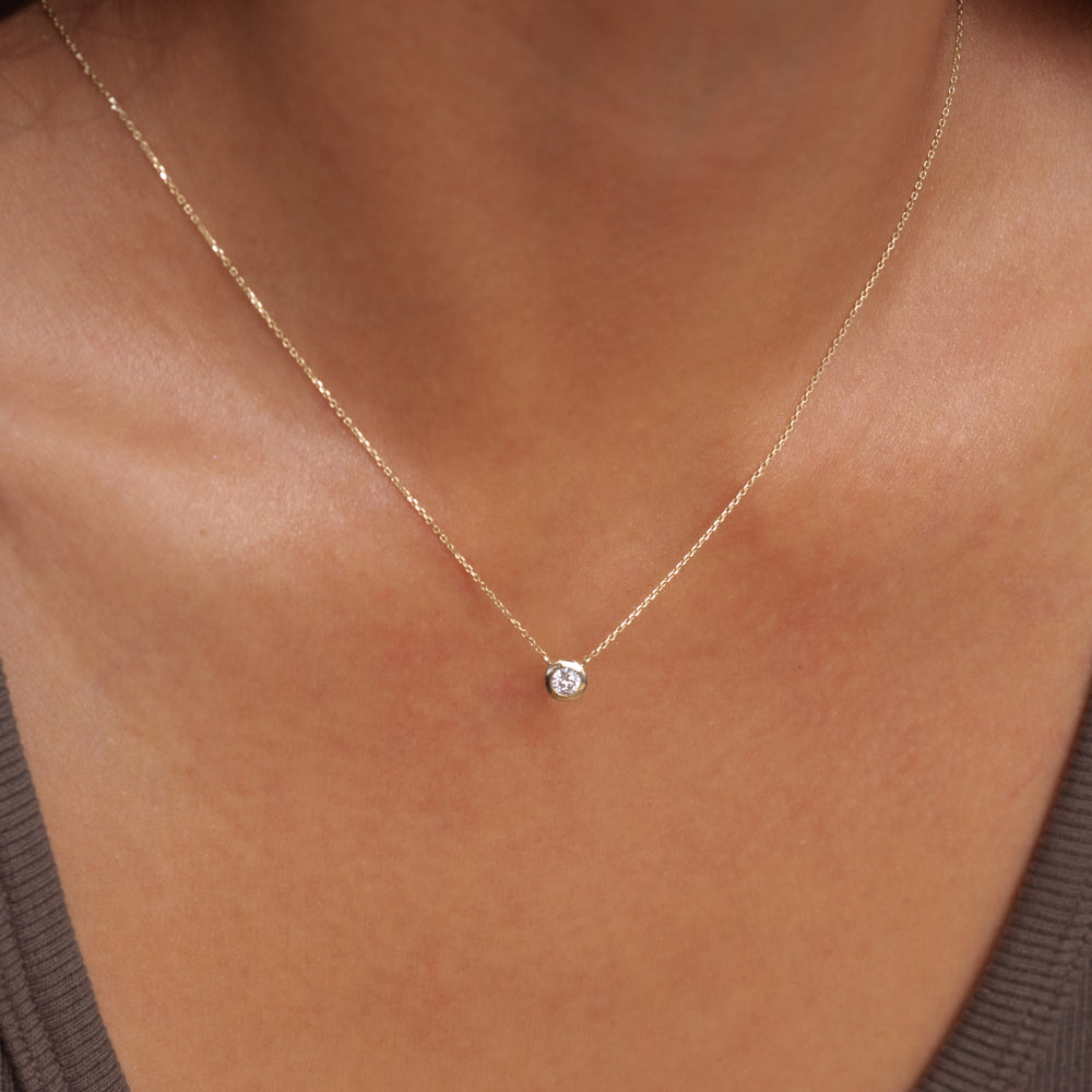 A delicate gold chain necklace with a small, clear round diamond pendant, shown worn on a person's neck.