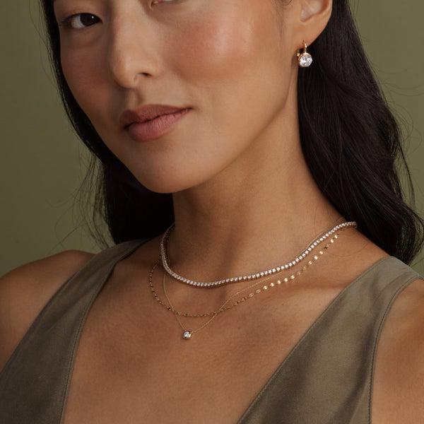 Close-up of a woman wearing layered delicate necklaces and a matching stud earring. The jewelry includes a short tennis chain, a fine gold chain, and a thin gold chain with a small clear gemstone pendant.