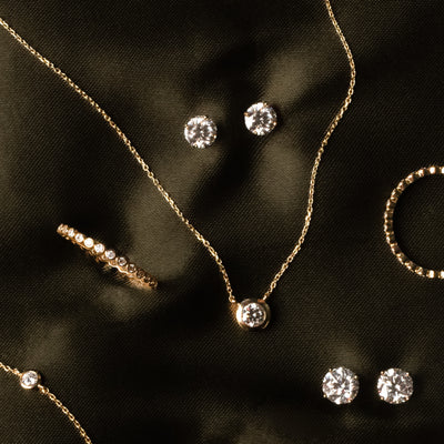 Gold jewelry set with a pendant necklace, ring, bracelet, and stud earrings, featuring clear diamonds on a dark satin fabric background.
