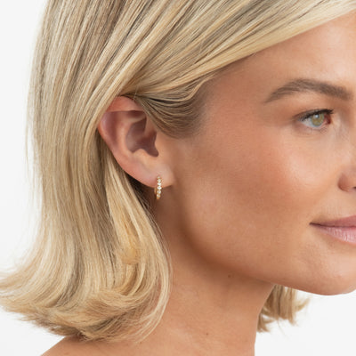 A close-up side profile of a woman with blonde hair wearing a small gold hoop earring with tiny clear stones, set against a clean white background.