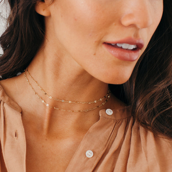 A close-up lifestyle shot of a woman wearing a delicate double-layered gold chain necklace with elongated oval links, styled with a tan button-up shirt.