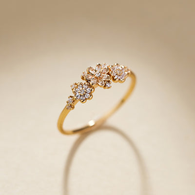 A gold ring featuring three small flower clusters, each set with clear stones, photographed on a warm beige background with a soft reflection below.