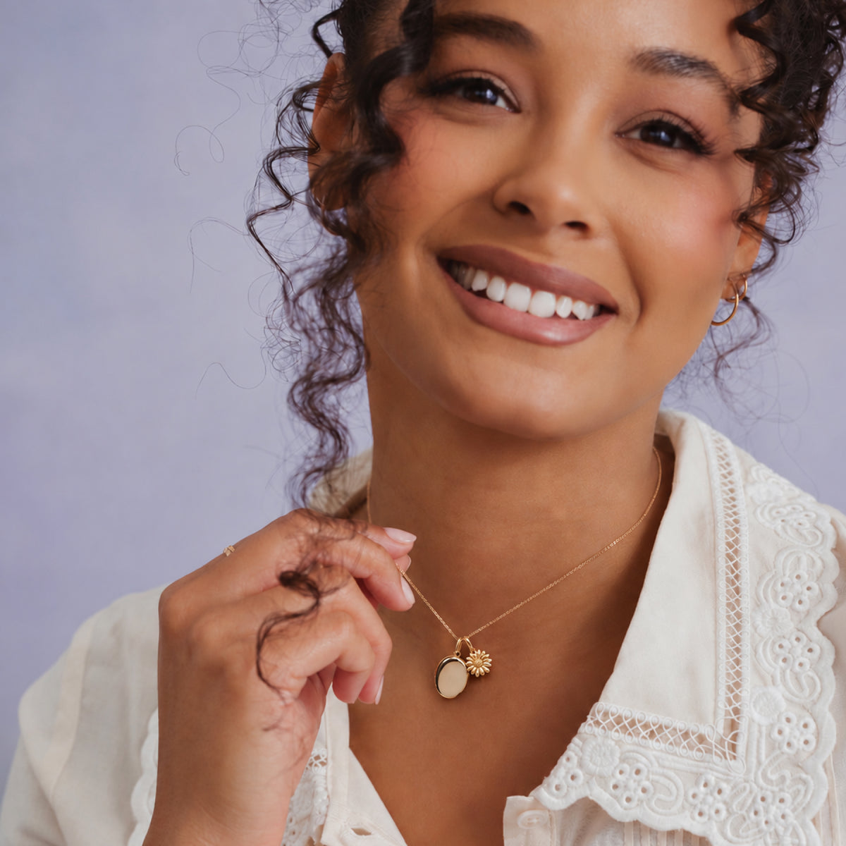 A close-up lifestyle shot of a smiling woman with curly hair wearing a delicate gold necklace with a flower and oval locket pendant, paired with a white lace collar blouse against a soft lavender background.